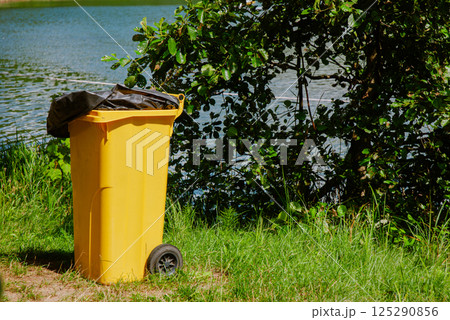 Bright yellow trash bin near lake in lush green park setting. Weekend concept near the forest and lake 125290856