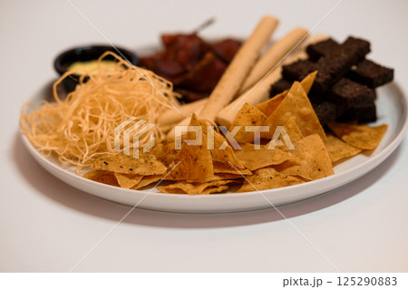 Assorted snack plate with tortilla chips, beef jerky, breadsticks, string cheese and dark chocolate cubes on a white background 125290883