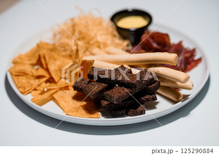 Variety snack plate with tortilla chips, cheese strings, dried meat, breadsticks and creamy dip on white background 125290884