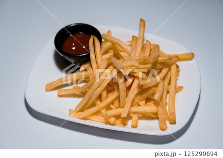Golden crispy french fries served on a white rectangular plate with a small black bowl of ketchup sauce, isolated on light background 125290894
