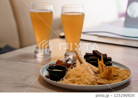 Two glasses of golden beer are positioned next to a plate filled with snacks like nachos, pretzels, and dark chocolate sticks on a light wooden table 125290920