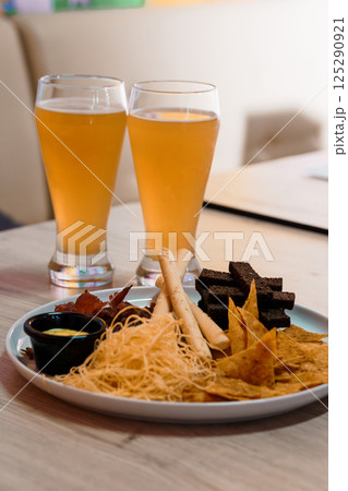 Two glasses of amber beer stand beside a plate containing tortilla chips, breadsticks, chocolate squares and a small dish with yellow sauce on wooden table 125290921