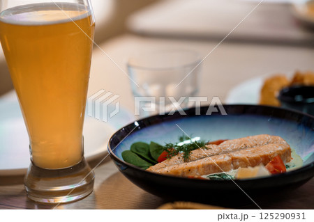Close-up of a glass of beer alongside a plate with salmon, spinach and tomatoes on a table in a restaurant setting Close-up of a glass of beer alongside a plate with salmon, spinach and tomatoes on a table in a restaurant setting 125290931