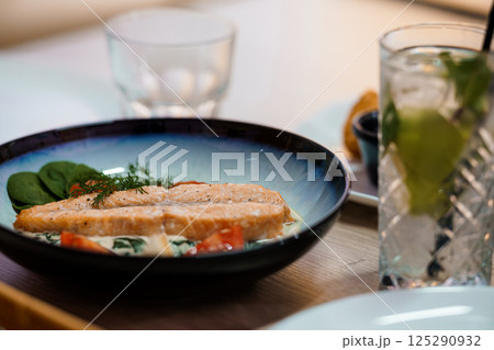Delicious salmon fillet served on a blue plate with spinach, tomatoes and dill, alongside a refreshing drink in a restaurant setting 125290932