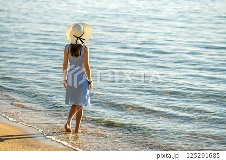 Young woman in straw hat and a dress standing alone on empty sand beach at sea shore. Lonely tourist girl looking at horizon over calm ocean surface on vacation trip. Young woman in straw hat and a dress standing alone on empty sand beach at sea shore. Lonely tourist girl looking at horizon over calm ocean surface on vacation trip. 125291685