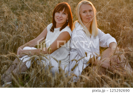 Two women sitting together in a golden wheat field during sunset sharing a moment of friendship and connection Two women sitting together in a golden wheat field during sunset sharing a moment of friendship and connection 125292690