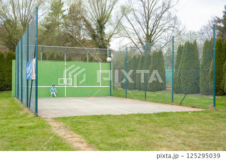Child in sports outfit plays with a ball at a green-walled court. Natural setting, solo action, and clear space perfect for campaigns on childhood development, sports safety, or active lifestyle. 125295039