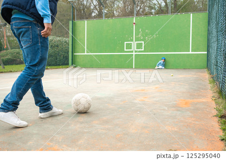 A quiet scene on a fenced playground. A child in blue sits safely in the corner while a man approaches a white ball. A calm yet dynamic family moment unfolds. A quiet scene on a fenced playground. A child in blue sits safely in the corner while a man approaches a white ball. A calm yet dynamic family moment unfolds. 125295040