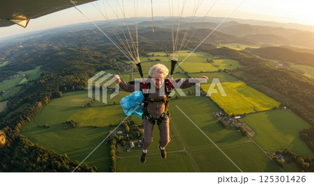 An elderly woman experiences the excitement of skydiving as she leaps from an aircraft, surrounded by stunning views of expansive green fields and hills. The sun sets in the background, casting a warm 125301426