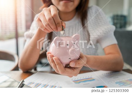 Close-up of a woman inserting a coin into a pink piggy bank, symbolizing savings and financial planning. Close-up of a woman inserting a coin into a pink piggy bank, symbolizing savings and financial planning. 125303300