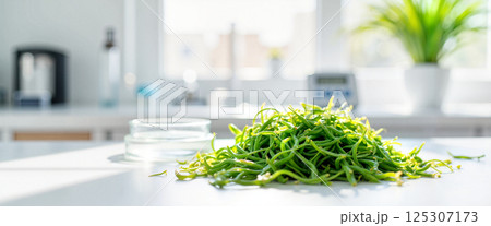 Fresh green vegetable pile sits on a table, surrounded by bright natural light from windows in a modern kitchen. A glass dish is nearby, and a potted plant is visible in the background 125307173
