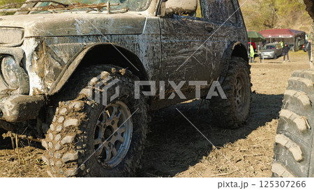 A close-up of a muddy off-road vehicle with large, knobby tires. The vehicle is covered in mud. Other vehicles and spectators are visible in the background A close-up of a muddy off-road vehicle with large, knobby tires. The vehicle is covered in mud. Other vehicles and spectators are visible in the background 125307266