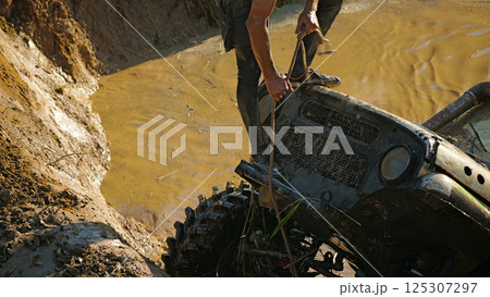 A male crew navigator stands on the hood of an SUV stuck in a muddy ditch and pulls the winch rope. Extreme racing 125307297