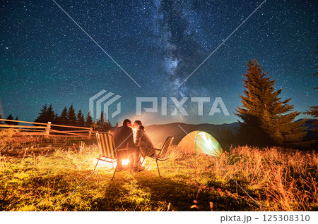 Couple hikers shares kiss by campfire, seated in chairs under magnificent starry sky with Milky Way visible above. Illuminated tent and serene mountain backdrop enhance romantic, tranquil ambiance. 125308310