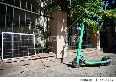 Mint-green electric scooter parked on stone pavement next to solar panel leaning against iron fence. Solar panel connected to scooter, emphasizing sustainable method of charging in urban setting. 125308313