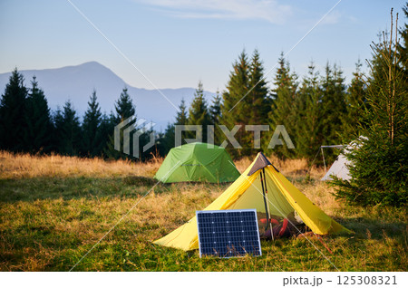 Solar panel in grassy field with tents in background highlights eco-friendly camping setup, surrounded by dense evergreen trees in evening. Integration of renewable energy in outdoor activities. 125308321