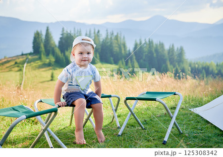 Young boy sits barefoot on small folding stool in grassy area, with two empty stools beside him. He wears white cap backwards and patterned shirt. Lush, green hills and mountains under clear sky. 125308342