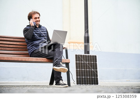 Man sits on wooden bench, working on laptop and smartphone, both connected to solar panel. Urban setting and casual attire highlight modern, sustainable, mobile workspace. Man sits on wooden bench, working on laptop and smartphone, both connected to solar panel. Urban setting and casual attire highlight modern, sustainable, mobile workspace. 125308359