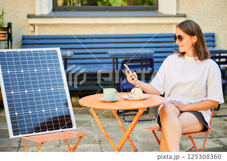 Young woman charging her smartphone from solar panel while relaxing. Pretty girl drinking coffee and checking her phone next to solar battery. Conscious female choosing sustainable lifestyle. Young woman charging her smartphone from solar panel while relaxing. Pretty girl drinking coffee and checking her phone next to solar battery. Conscious female choosing sustainable lifestyle. 125308360