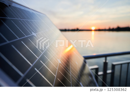 Solar panel reflects setting sun over tranquil lake, with railing in background. Integration of clean energy technology with natural beauty, showcasing harmonious blend of innovation and environment. Solar panel reflects setting sun over tranquil lake, with railing in background. Integration of clean energy technology with natural beauty, showcasing harmonious blend of innovation and environment. 125308362