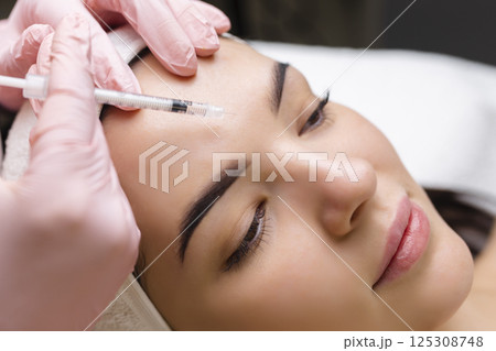 Close up of hands of young cosmetologist injecting botox in female face. She is standing and smiling. The woman is closed her eyes with relaxation 125308748