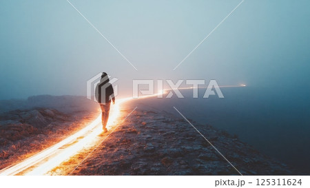 A solitary figure strolls along a rocky path, shrouded in mist and twilight. Subtle glowing lights stretch along the ground, creating an enchanting trail that guides the way. The atmosphere is calm 125311624