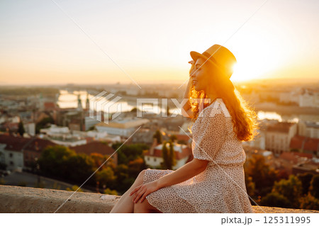 Smiling female tourist in hat admires landscape of city at dawn. Euro-trip. Active lifestyle concept 125311995