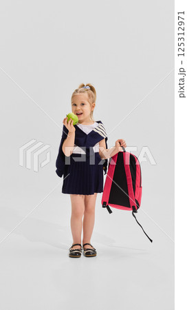 Smiling girl in navy pleated uniform eating green apple while holding backpack in one hand against white background. 125312971