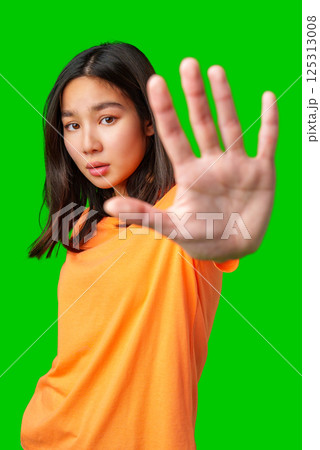 Young woman in orange shirt poses with hand raised against bright green background 125313008