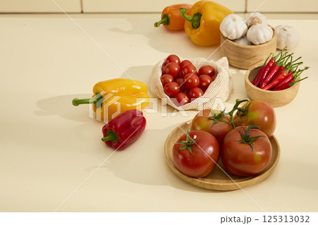 Tomatoes on a round wooden plate next to a bag of cherry tomatoes. Wooden bowls containing garlic and chili peppers. Fresh bell peppers with shiny peels. Shadows of objects cast on the kitchen table. 125313032