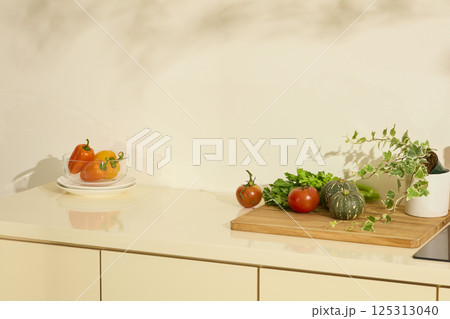 A glass bowl of bright yellow bell peppers sits on a stack of white porcelain plates. Fresh vegetables sit on a wooden cutting board, ready to be processed into a dish. Photo for product displaying. 125313040