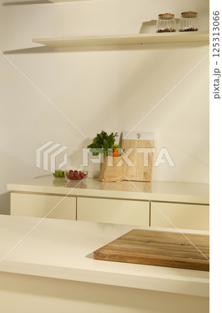 Vertical photo of the kitchen room. Vegetables in paper bags, next to glass bowls of tomatoes and green grapes. Empty wooden cutting board placed on white table in the middle of the kitchen 125313066