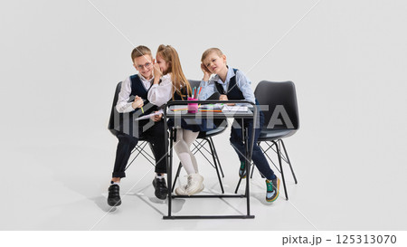 Girl whispering into boy's ear while seated at shared desk with pencils and notebooks as third classmate reacts. Concept of school gossip, 125313070