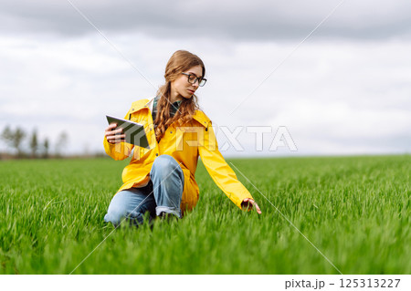 Young farmer woman in a green field, intently using a tablet. She wears a yellow jacket and glasses Young farmer woman in a green field, intently using a tablet. She wears a yellow jacket and glasses 125313227