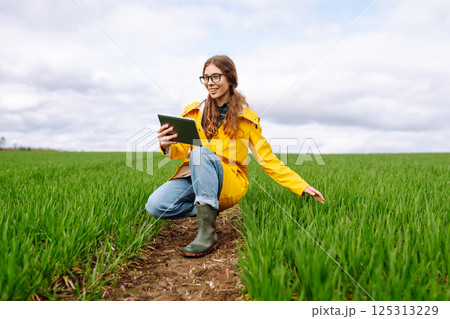 Young farmer woman in a green field, intently using a tablet. She wears a yellow jacket and glasses 125313229