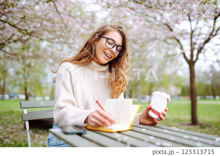 Happy woman relaxing in park, writing in notebook, sitting with laptop. Weekend, spring concept. Happy woman relaxing in park, writing in notebook, sitting with laptop. Weekend, spring concept. 125313715