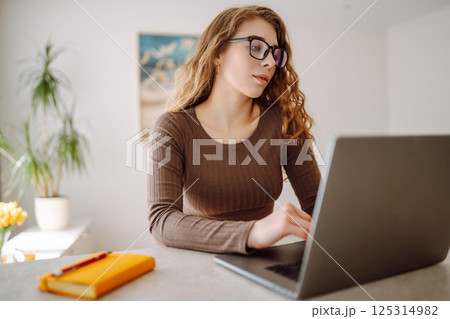Young woman uses laptop at home, looks at screen, reads or writes at table. The concept of education 125314982