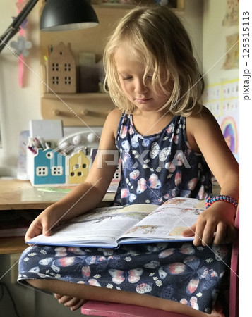 Cute girl with blond hair reading a book sitting cross-legged on a chair Cute girl with blond hair reading a book sitting cross-legged on a chair 125315113