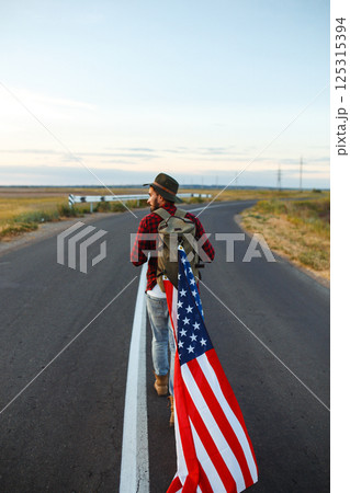4th of July. American Flag. Traveler with the flag of America. The man in a hat, a backpack, a shirt and jeans. 4th of July. American Flag. Traveler with the flag of America. The man in a hat, a backpack, a shirt and jeans. 125315394