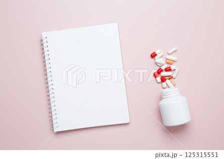 White bottle container with colorful medication capsules and blank notebook. Concept of medicine, healthcare, and pharmaceutical industry 125315551