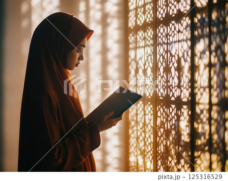 Woman in a hijab praying at home, holding a misbaha, with soft light through patterned windows 125316529