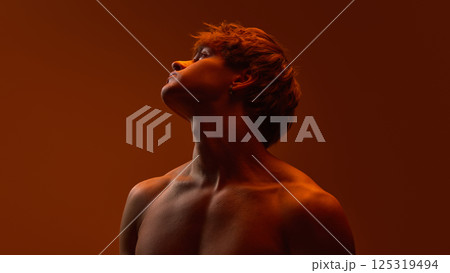 Horizontal portrait of young man with intense gaze lifting head in deep orange shadowed light with sculpted shoulders. 125319494