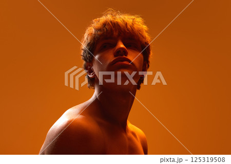 Shirtless young man gazing upward with serious expression in glowing orange light against gradient studio background. 125319508