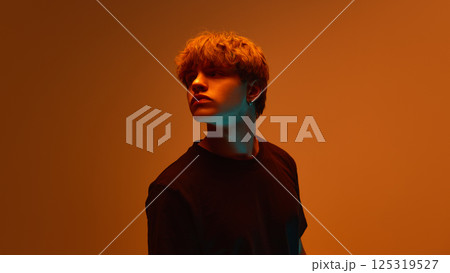 Young man turning head to side, with calm and focused expression, posing under strong contrast lighting under orange hue. 125319527