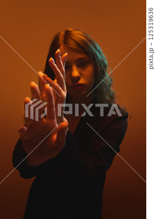 Portrait of young focused woman reaching forward with expressive gesture under bright warm orange light against gradient background. 125319636