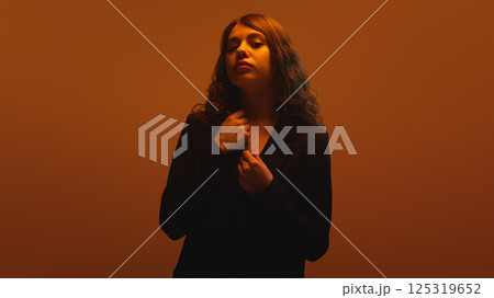 Portrait of young woman in black outfit holding collar, gazing with calm defiance against orange backdrop in neon light. Concept of introspection Portrait of young woman in black outfit holding collar, gazing with calm defiance against orange backdrop in neon light. Concept of introspection 125319652