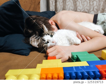 A cheerful young boy hugging his white and gray cat while lying on the couch, portraying companionship and love. happy boy cuddles with his friend the cat A cheerful young boy hugging his white and gray cat while lying on the couch, portraying companionship and love. happy boy cuddles with his friend the cat 125319713