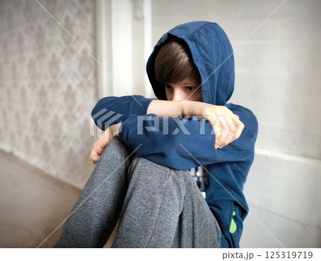 sad young man is sitting on the floor. A depressed teenager in a hood is sitting on the floor in the room. 125319719