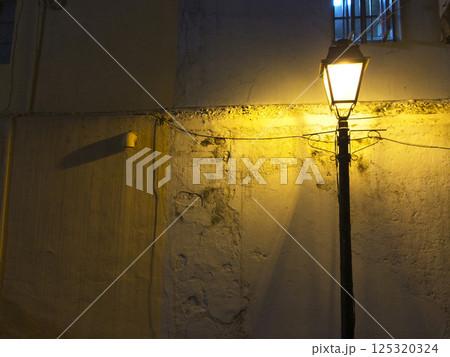 夜の路地裏の黒い街灯が照らす温かいオレンジ色の光の中の古びた剥がれかけた表面の白い壁と鉄格子のある窓 夜の路地裏の黒い街灯が照らす温かいオレンジ色の光の中の古びた剥がれかけた表面の白い壁と鉄格子のある窓 125320324