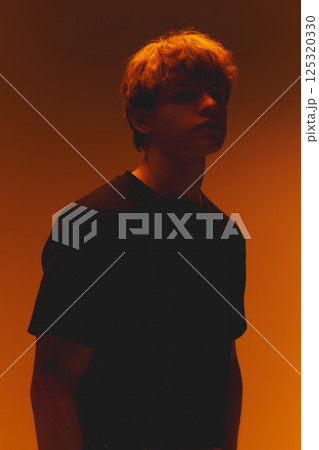 Young man in shadows, face partially hidden, in warm dramatic neon lighting against warm orange gradient background. Concept of beauty 125320330